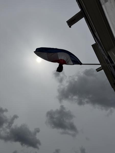 The photo captures a Dutch flag, silhouetted against a dramatic cloudy sky, with the sun peeking through the dense clouds